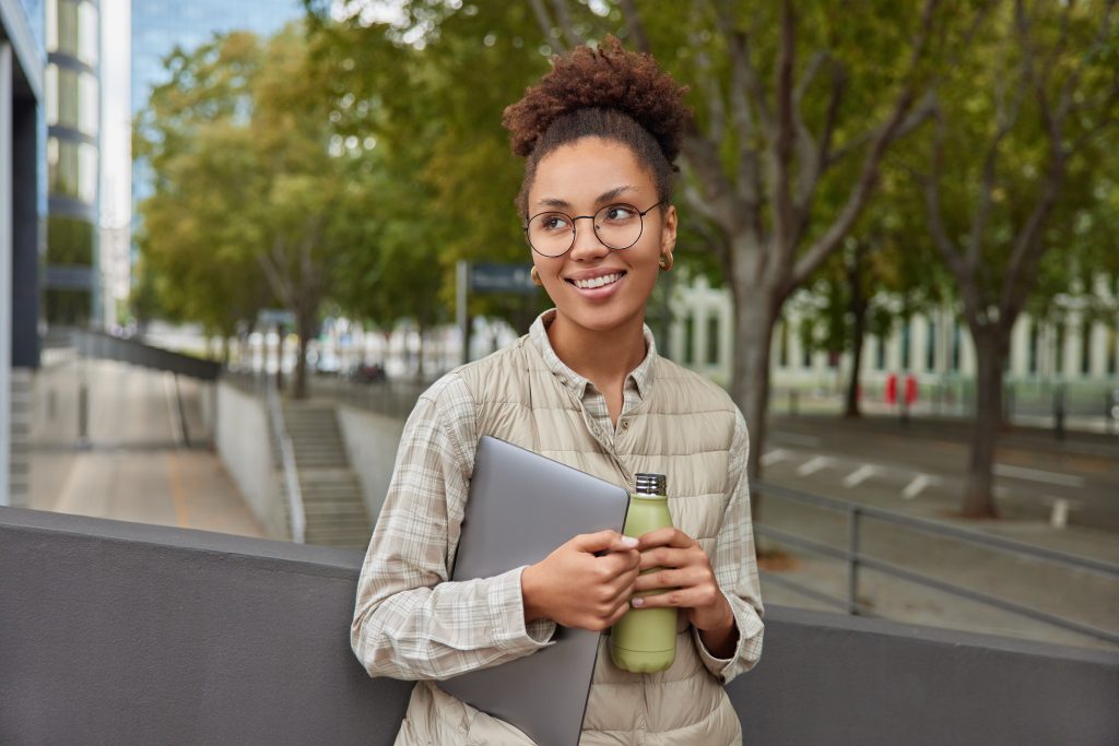 Jovem mulher com óculos redondos e cabelo cacheado preso em um coque segura um notebook e uma garrafa reutilizável em ambiente urbano arborizado, sorrindo enquanto observa ao redor.