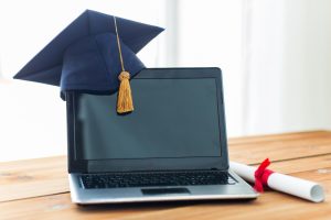 Laptop sobre uma mesa de madeira com um capelo de formatura apoiado na tela e um diploma enrolado ao lado.