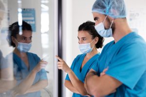 Dois profissionais de saúde, um homem e uma mulher, vestindo uniformes azuis e máscaras, discutem frente a um vidro refletivo em um ambiente hospitalar.